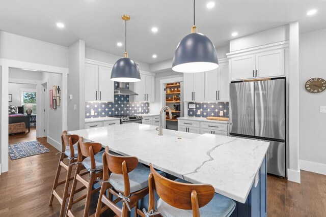 kitchen featuring wall chimney range hood, appliances with stainless steel finishes, a center island with sink, a sink, and white cabinets