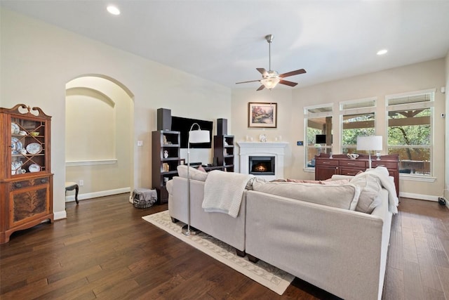 living room with a warm lit fireplace, ceiling fan, dark wood-style floors, and baseboards