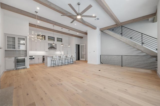 unfurnished living room with light wood-type flooring, beam ceiling, ceiling fan with notable chandelier, wine cooler, and recessed lighting