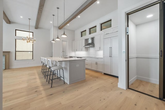 kitchen featuring a kitchen breakfast bar, a center island with sink, baseboards, white cabinets, and wall chimney exhaust hood