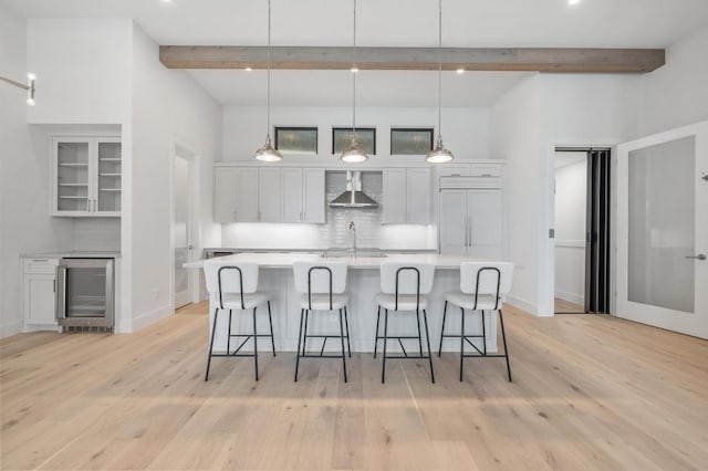 kitchen featuring wall chimney range hood, beam ceiling, white cabinetry, wine cooler, and backsplash