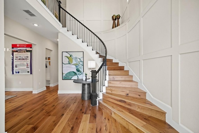 stairs featuring a decorative wall, wood-type flooring, baseboards, a towering ceiling, and visible vents