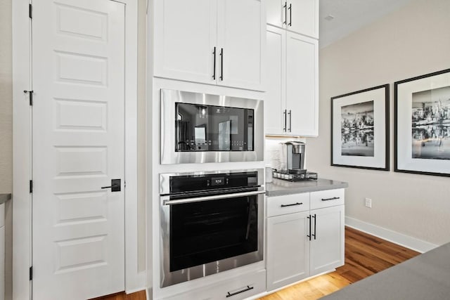 kitchen featuring stainless steel oven, built in microwave, white cabinetry, baseboards, and light wood-type flooring