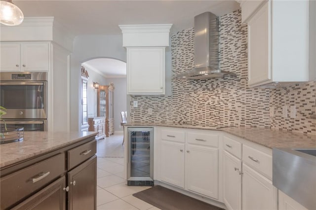 kitchen featuring wall chimney range hood, backsplash, arched walkways, double oven, and beverage cooler