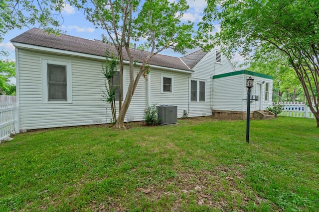 rear view of property featuring a lawn, roof with shingles, central air condition unit, and fence