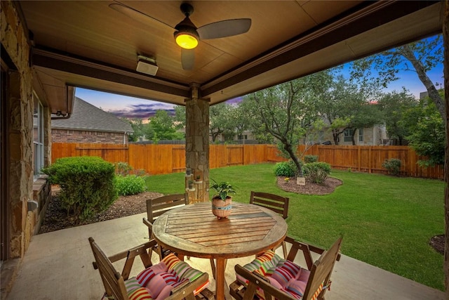 patio terrace at dusk with outdoor dining area, a fenced backyard, ceiling fan, and a lawn