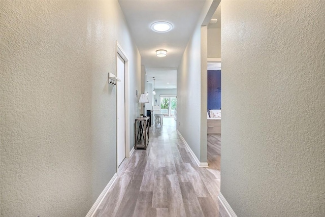 hallway featuring baseboards, a textured wall, and light wood-type flooring