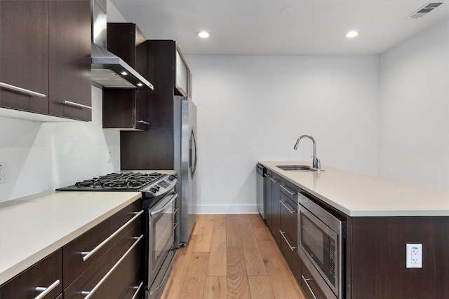 kitchen featuring wall chimney range hood, appliances with stainless steel finishes, light wood-type flooring, dark brown cabinets, and a sink