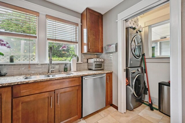 kitchen featuring dishwasher, stacked washer / drying machine, a sink, brown cabinetry, and tasteful backsplash