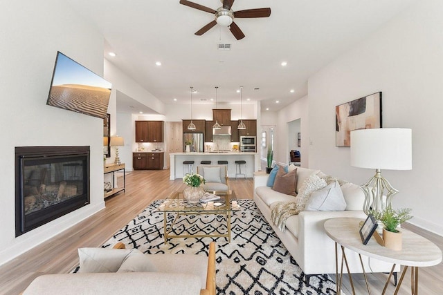 living room featuring a ceiling fan, visible vents, a glass covered fireplace, recessed lighting, and light wood-style flooring