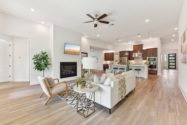 living room featuring light wood-style floors, a ceiling fan, a glass covered fireplace, and visible vents