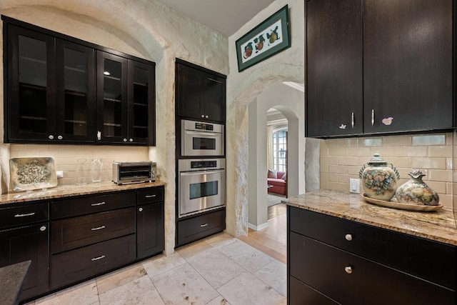 kitchen featuring arched walkways, tasteful backsplash, light stone countertops, a toaster, and dark cabinetry