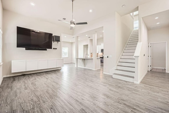 unfurnished living room featuring a ceiling fan, light wood-style floors, baseboards, stairway, and a sink