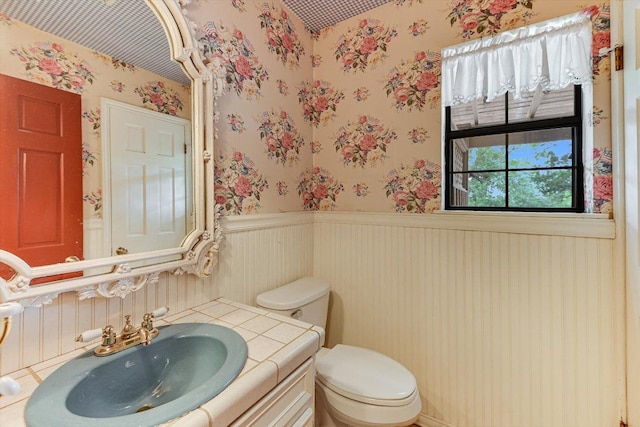 half bathroom featuring toilet, wallpapered walls, wainscoting, and vanity