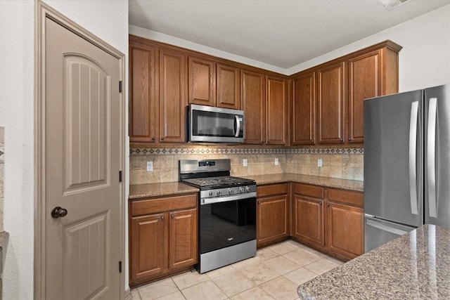 kitchen featuring brown cabinets, light stone countertops, stainless steel appliances, light tile patterned floors, and backsplash