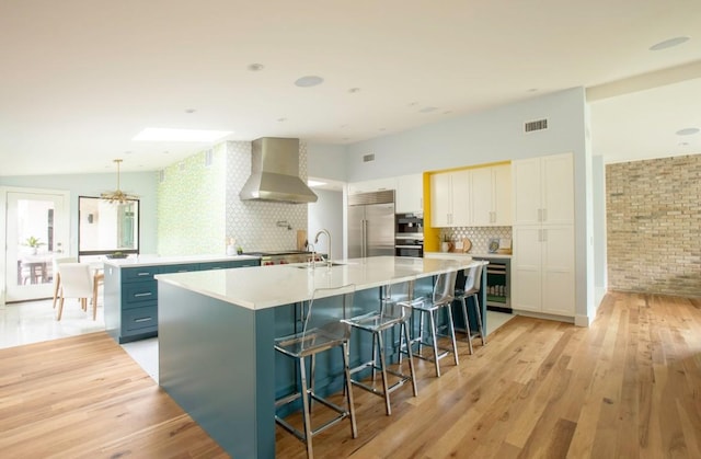 kitchen featuring wall chimney range hood, a sink, light wood-style flooring, stainless steel appliances, and a spacious island