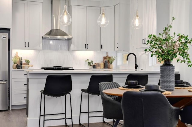 kitchen with decorative backsplash, a kitchen breakfast bar, light countertops, white cabinets, and wall chimney exhaust hood