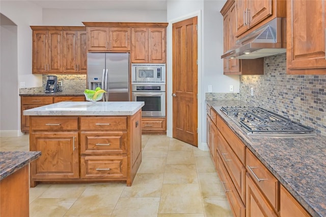 kitchen with brown cabinetry, decorative backsplash, under cabinet range hood, dark stone countertops, and appliances with stainless steel finishes