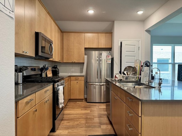kitchen with decorative backsplash, a kitchen island with sink, a sink, light wood-style flooring, and appliances with stainless steel finishes