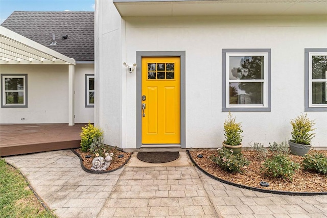 view of exterior entry featuring roof with shingles, stucco siding, and a deck