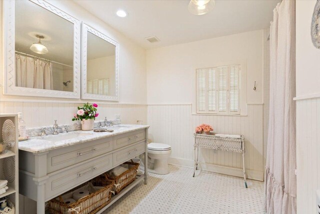 bathroom with toilet, a sink, tile patterned floors, double vanity, and a wainscoted wall