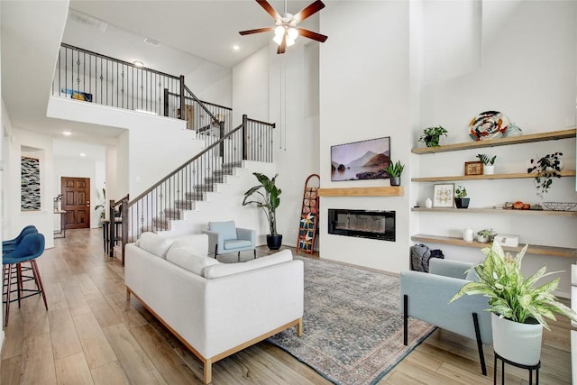 living area with a ceiling fan, a glass covered fireplace, a towering ceiling, wood finished floors, and stairway