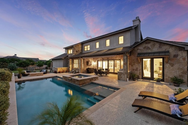 back of property at dusk featuring stone siding, a patio area, a pool with connected hot tub, and a chimney