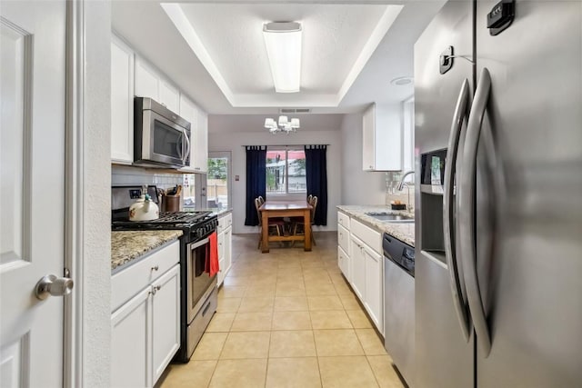 kitchen with a sink, stainless steel appliances, a notable chandelier, white cabinetry, and a raised ceiling