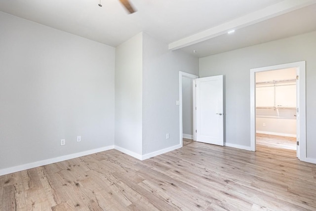 unfurnished bedroom featuring ceiling fan, light wood-style flooring, beamed ceiling, a walk in closet, and baseboards
