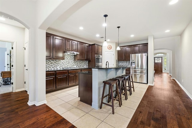 152 Florenz Ln, Georgetown, TX 78628 - kitchen with under cabinet range hood, light wood finished floors, a kitchen bar, stainless steel fridge, and dark countertops