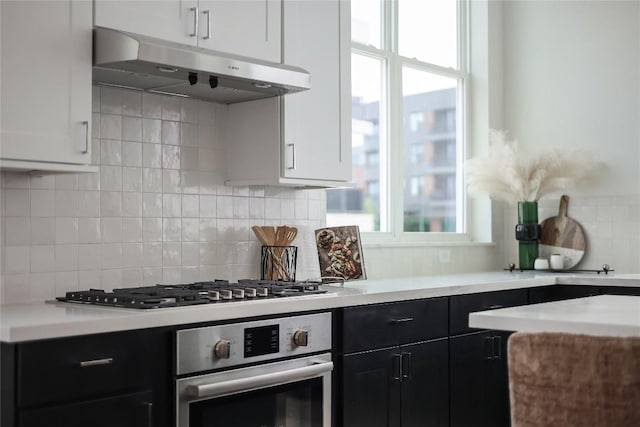 kitchen with dark cabinets, under cabinet range hood, light countertops, appliances with stainless steel finishes, and tasteful backsplash