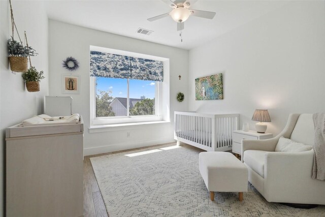 bedroom featuring wood finished floors, a nursery area, visible vents, baseboards, and ceiling fan
