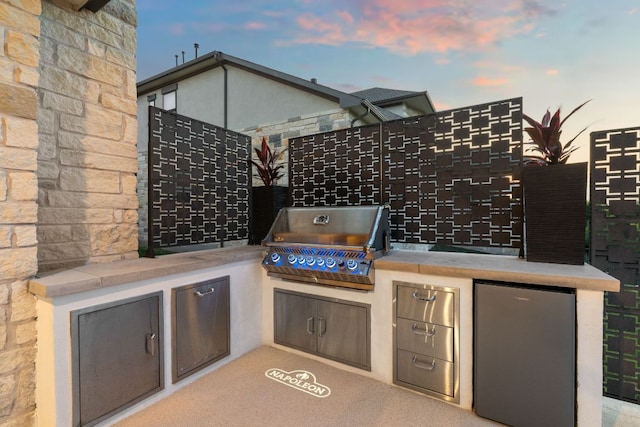 patio terrace at dusk with fence, a grill, and an outdoor kitchen
