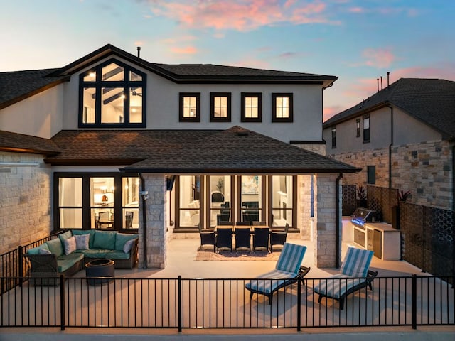 back of house at dusk featuring a patio area, stone siding, an outdoor living space, a shingled roof, and an outdoor kitchen