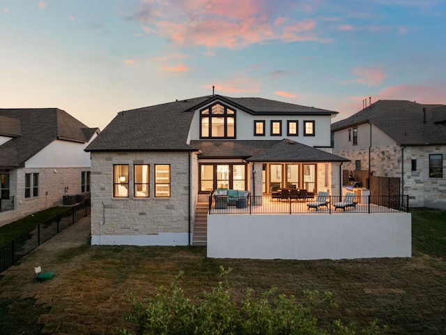 back of property at dusk with stone siding, roof with shingles, a lawn, and a fenced backyard