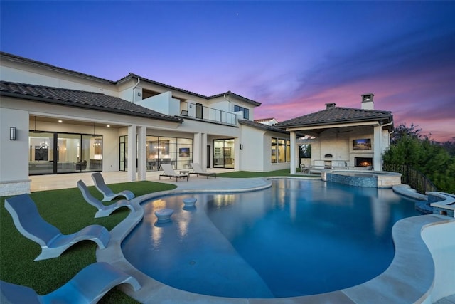 5609 Laceback Ter, Austin, TX 78738 - back of property at dusk featuring a balcony, a patio area, a pool with connected hot tub, a warm lit fireplace, and a tile roof