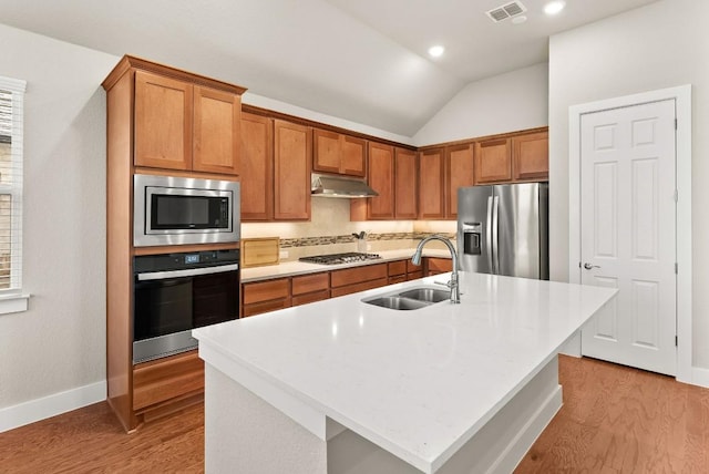 kitchen featuring appliances with stainless steel finishes, a sink, under cabinet range hood, vaulted ceiling, and brown cabinetry