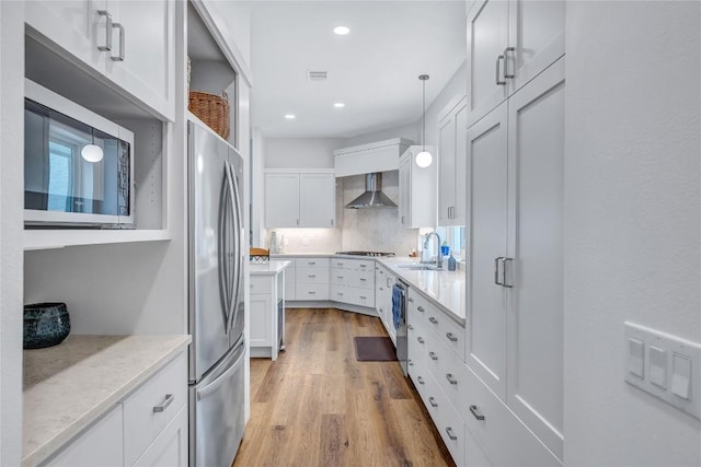 kitchen with appliances with stainless steel finishes, wall chimney range hood, light wood-style flooring, white cabinetry, and a sink