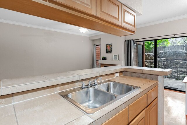 kitchen with a sink, tile counters, crown molding, light wood-style floors, and a ceiling fan