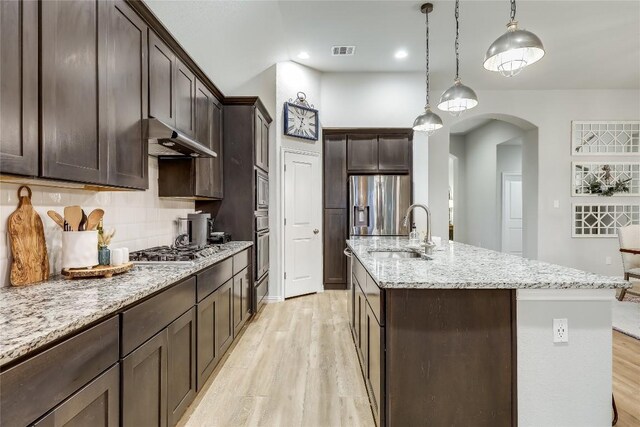 kitchen with under cabinet range hood, light wood-style floors, appliances with stainless steel finishes, dark brown cabinets, and a kitchen island with sink