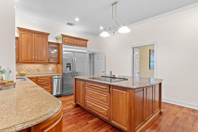 kitchen with built in refrigerator, black electric cooktop, an island with sink, wood finished floors, and crown molding