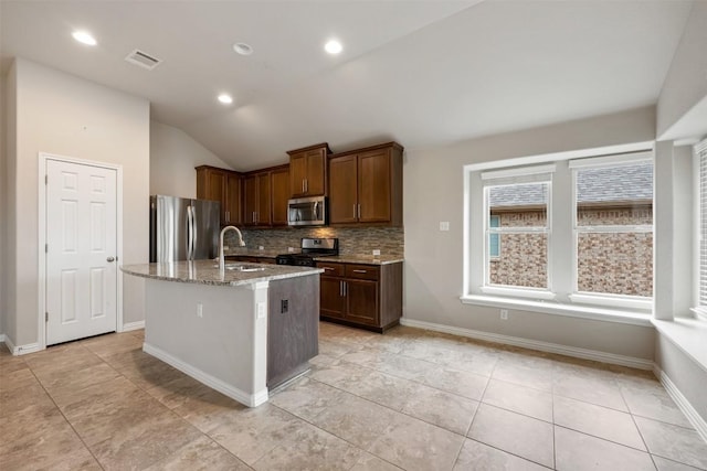 kitchen with appliances with stainless steel finishes, light stone counters, backsplash, an island with sink, and vaulted ceiling