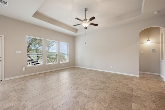 empty room with a raised ceiling, arched walkways, ceiling fan, light tile patterned floors, and baseboards