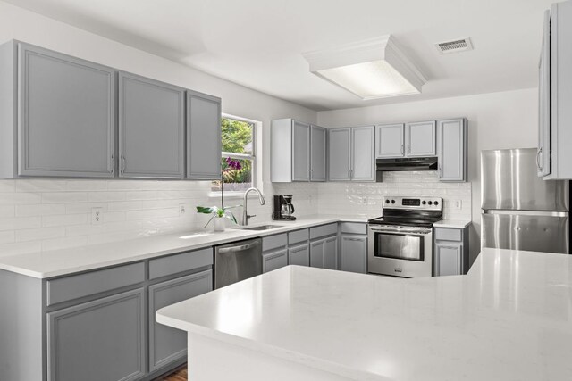 kitchen featuring gray cabinets, appliances with stainless steel finishes, a sink, under cabinet range hood, and decorative backsplash