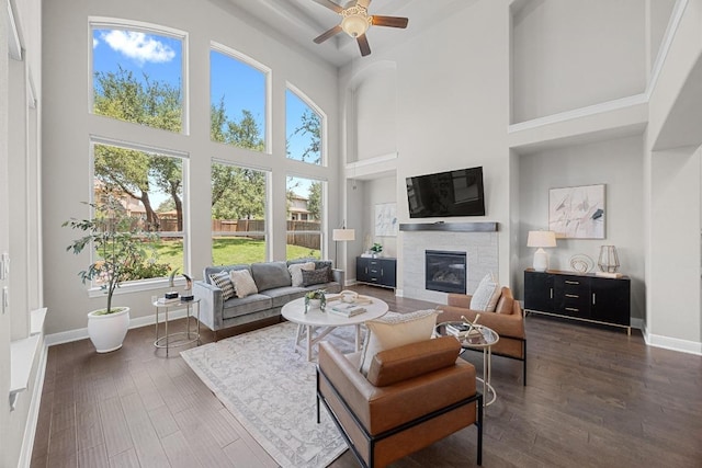 living area featuring a high ceiling, a glass covered fireplace, ceiling fan, dark wood-style floors, and baseboards