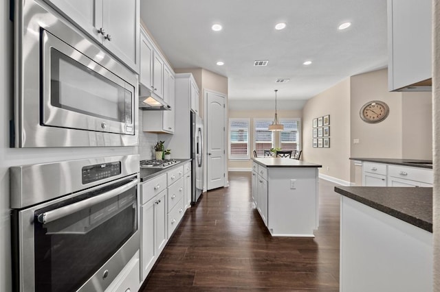 kitchen featuring stainless steel appliances, under cabinet range hood, a kitchen island, dark countertops, and dark wood-type flooring