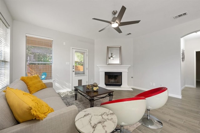 living room with arched walkways, wood finished floors, a fireplace with raised hearth, ceiling fan, and baseboards