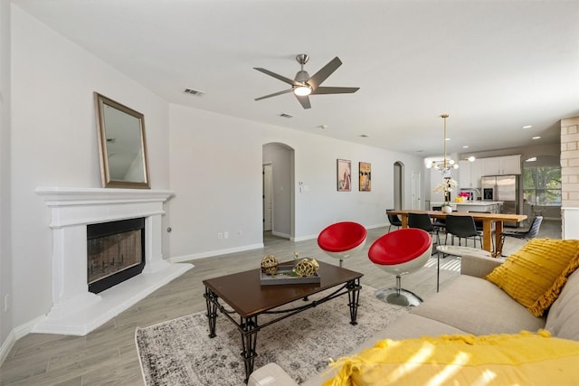 living area featuring arched walkways, ceiling fan, light wood-type flooring, a fireplace with raised hearth, and baseboards
