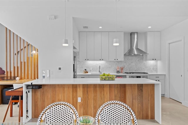 kitchen featuring wall chimney range hood, tasteful backsplash, light countertops, white cabinetry, and a peninsula