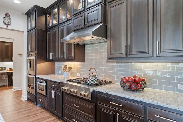 kitchen with under cabinet range hood, stainless steel appliances, light stone countertops, crown molding, and decorative backsplash